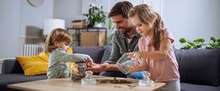 Ein Vater sitzt mit zwei kleinen Kindern am Wohnzimmertisch, während sie gemeinsam Münzen aus einem Glas zählen und sortieren – spielerische Vermittlung von Taschengeld, Geldwert und finanziellem Lernen im Familienalltag.