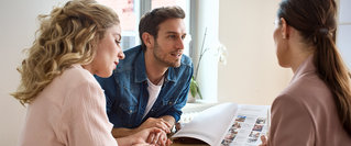 Ein Paar sitzt mit einer Finanzcoach an einem Tisch und bespricht Unterlagen – persönliche Beratung zum langfristigen Vermögensaufbau und zu finanzieller Planung.